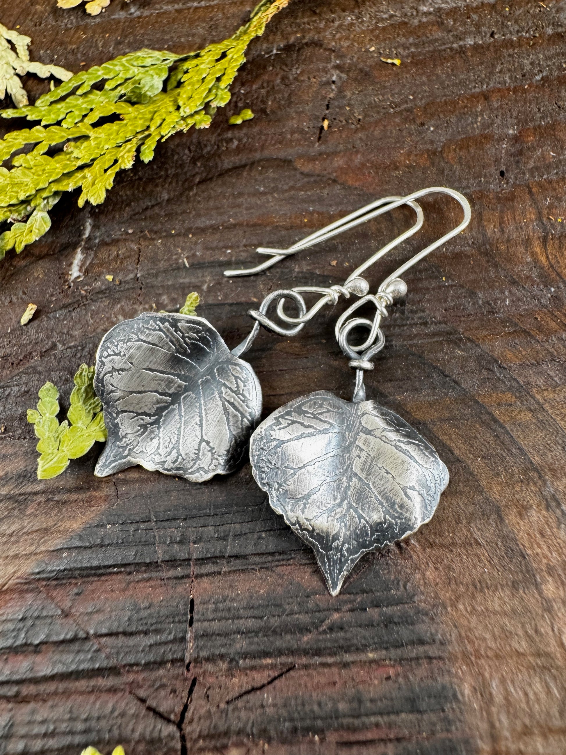 Silver leaf earrings on a wooden surface with green leaves.
