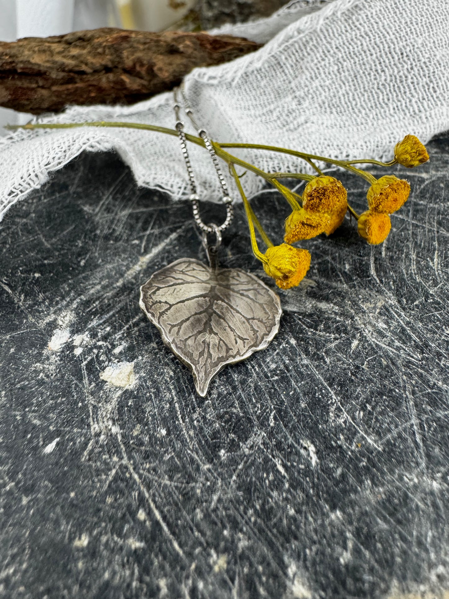 Silver leaf-shaped pendant on a chain with yellow flowers on a textured surface