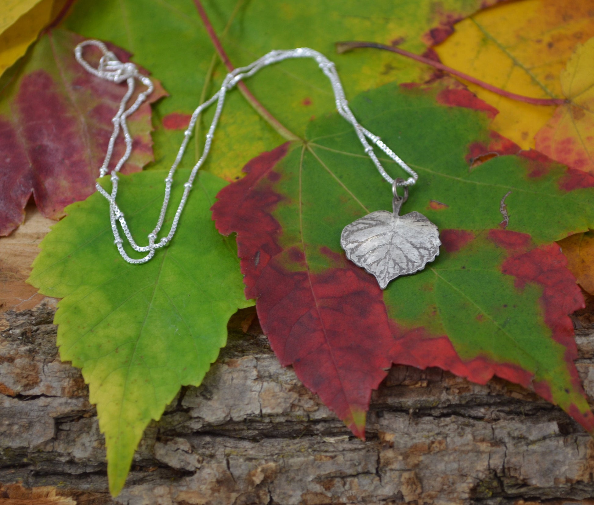 Birch Heart Leaf Necklace sitting on a colorful Maple Leaf and wood texture