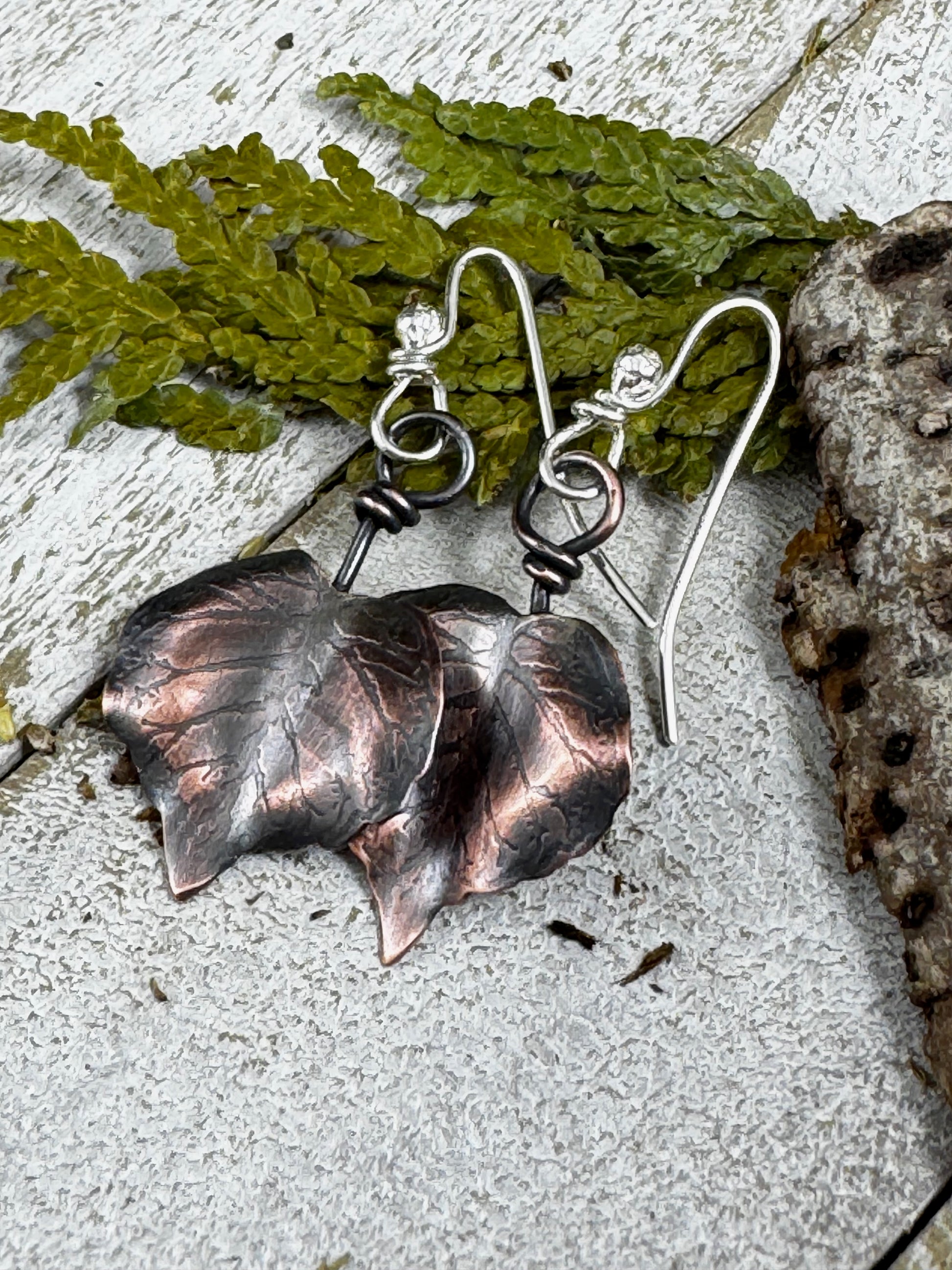 Copper leaf earrings on a textured surface with green leaves in the background