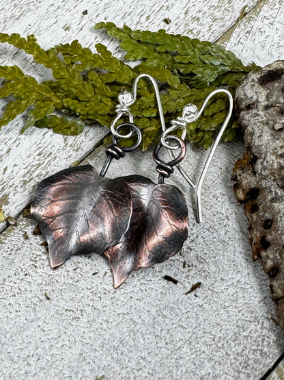 Copper leaf earrings on a textured surface with green leaves in the background