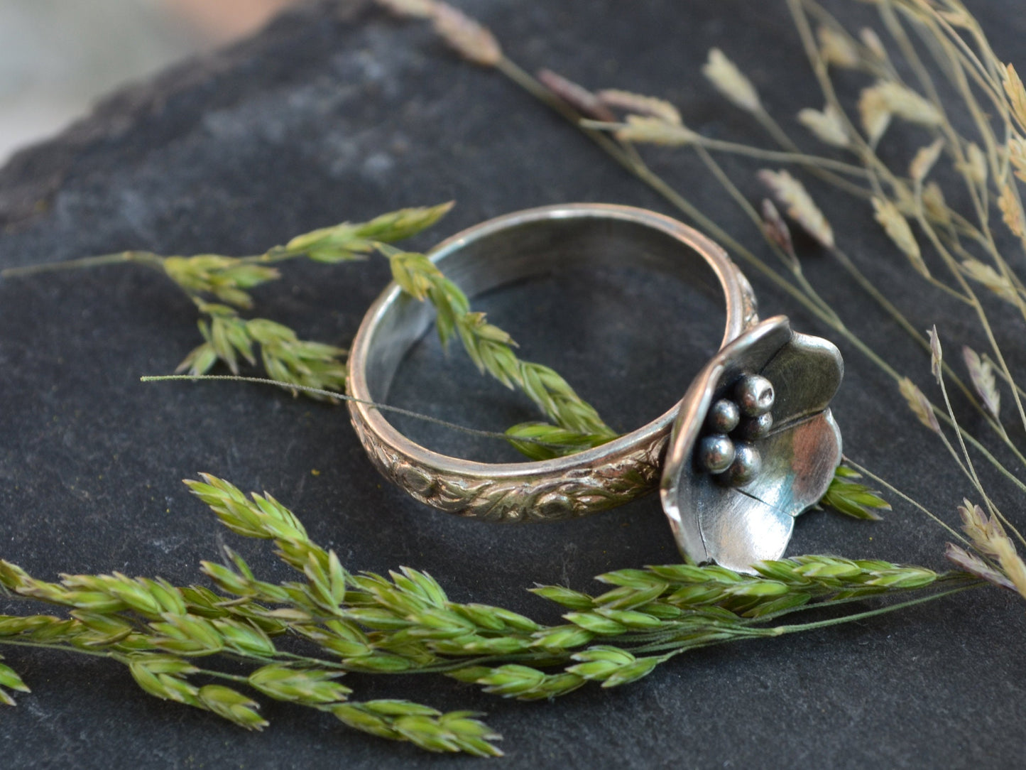 The Sterling Silver Deanna Flower Ring by Rust and Leaf displayed on a piece of slate with dried flowers